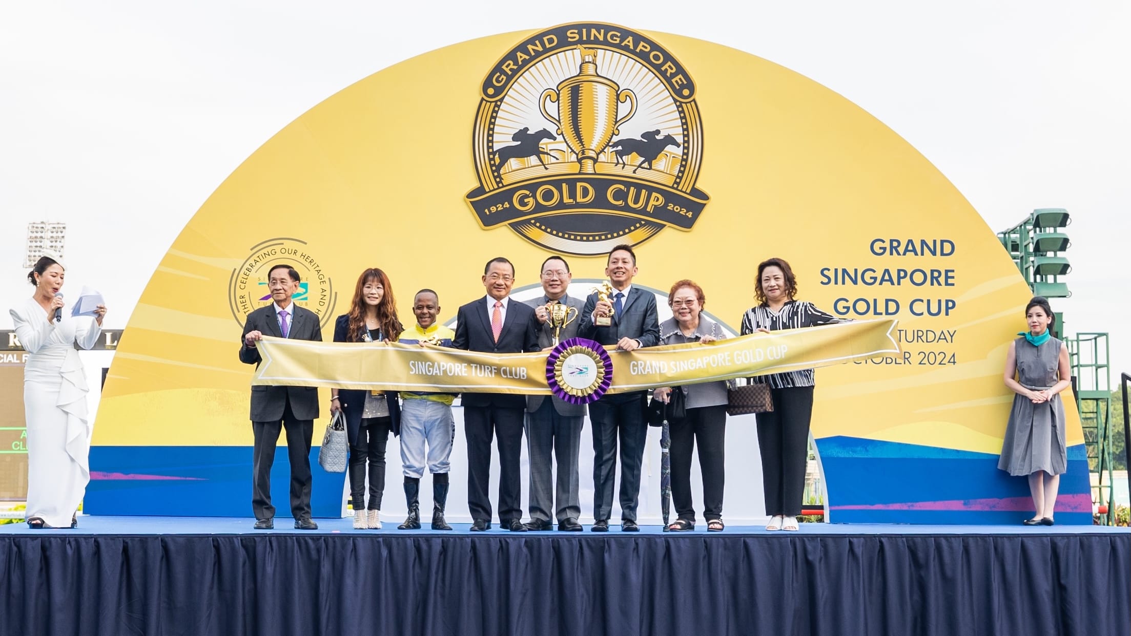 People on stage holding Singapore Turf Club banner; gold cup logo backdrop.